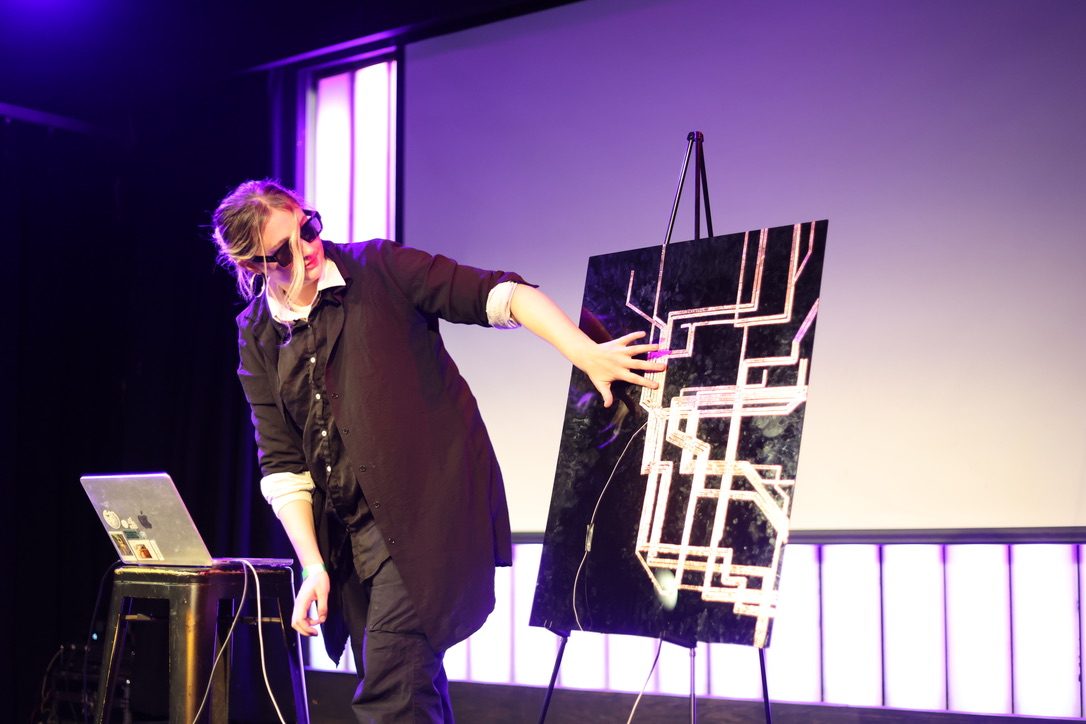 The artist on stage, hand on a copper-traced transit map on a black panel, beside a laptop.