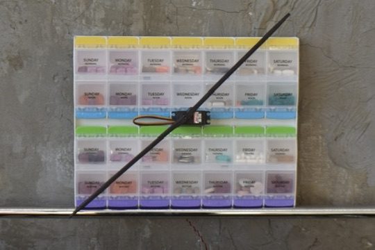 A pill organizer on a concrete wall with a clock hand across it.